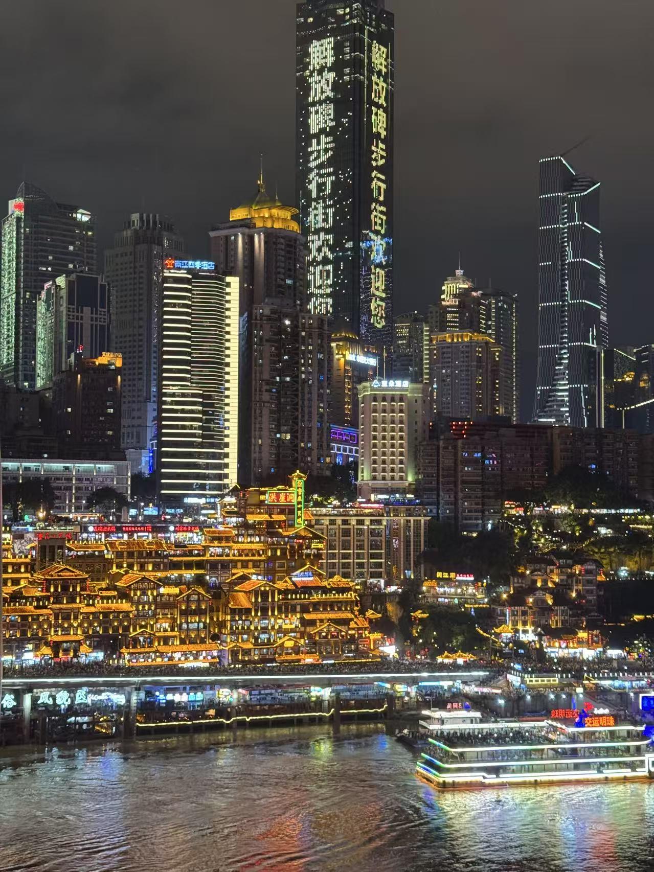 Chongqing at night: the golden stilt houses of Hongya Cave on the riverbank, lit towers rising behind them, and a boat crossing the water